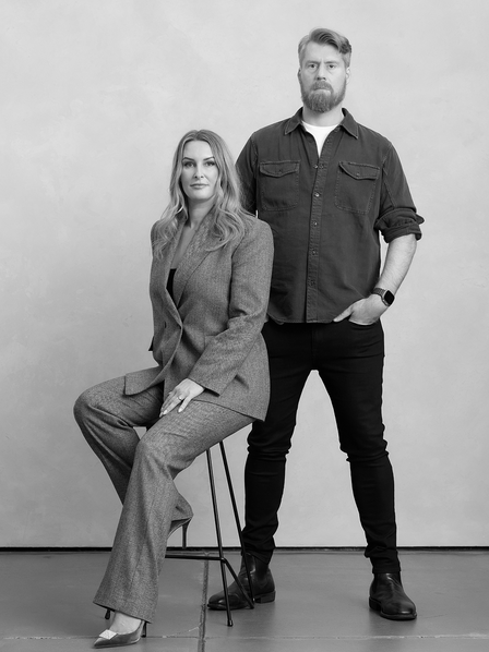 Black and white photo of a woman sitting on a chair and a man standing next to her against a plain background