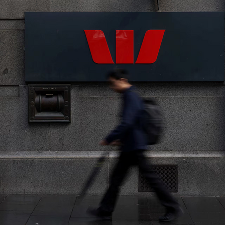 Person walking past a Westpac bank branch with a red logo on a gray wall.