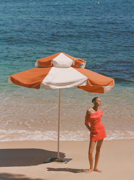 Woman in a red dress standing next to a large orange and white umbrella on a beach.