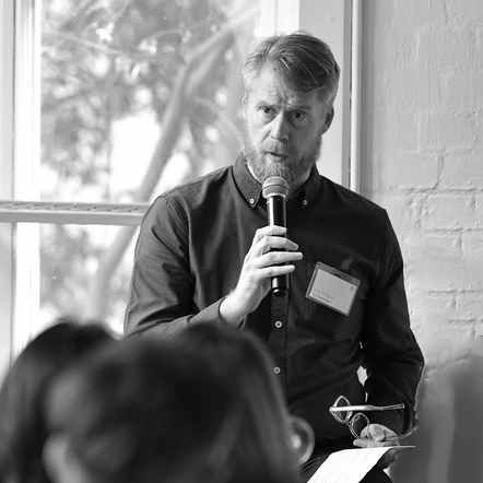Man speaking into a microphone at an event with blurred audience in foreground