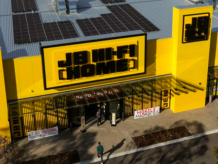 Aerial view of JB Hi-Fi retail store exterior with iconic yellow and black signage, solar panels on roof, and promotional banners at entrance