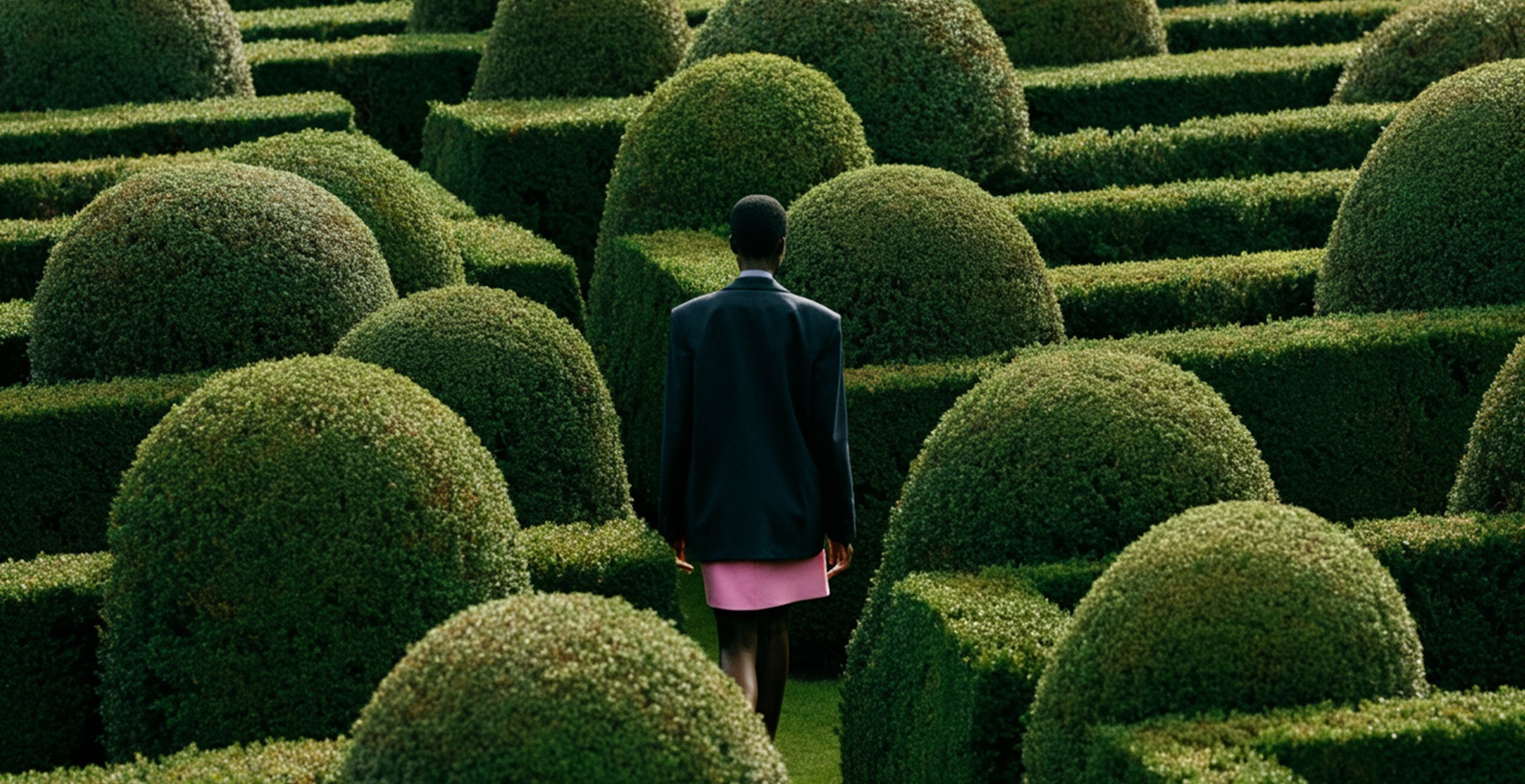 Woman walking through a hedge maze, wearing a black leather blazer and pink mini skirt. 