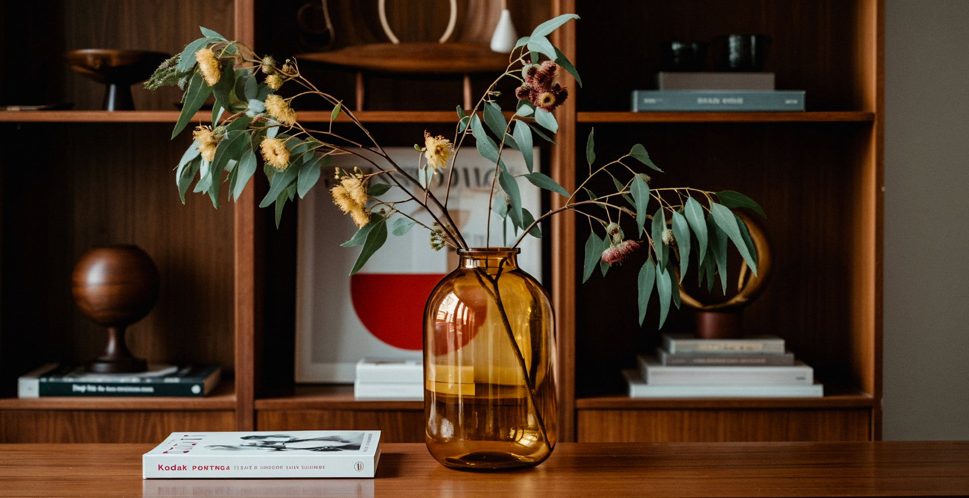 Detail shot of an amber coloured vase with shelving behind it featuring books and decorative objects.