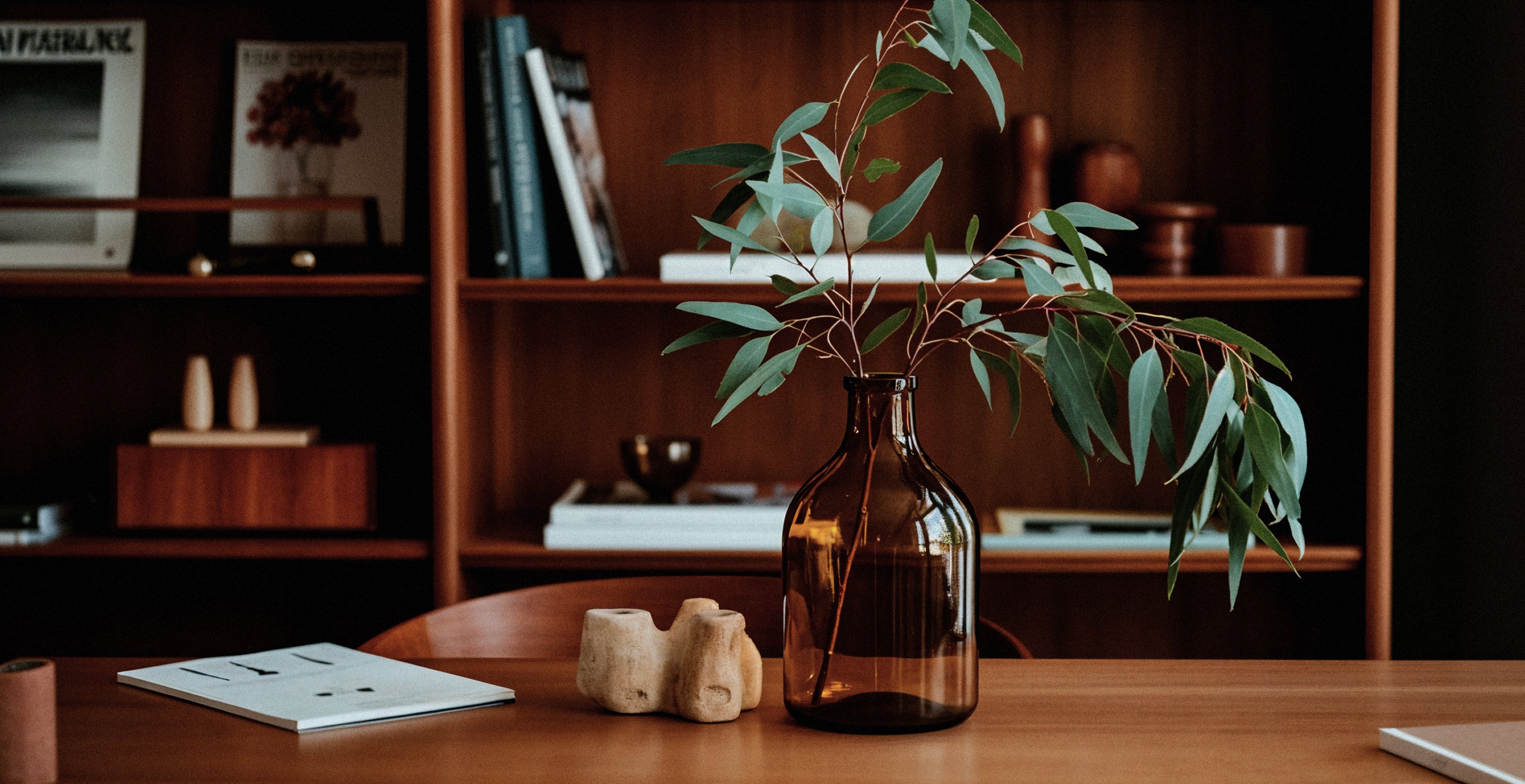Amber coloured vase with Eucalyptus leaves on a table, with shelving featuring books and decorative objects in the background. 