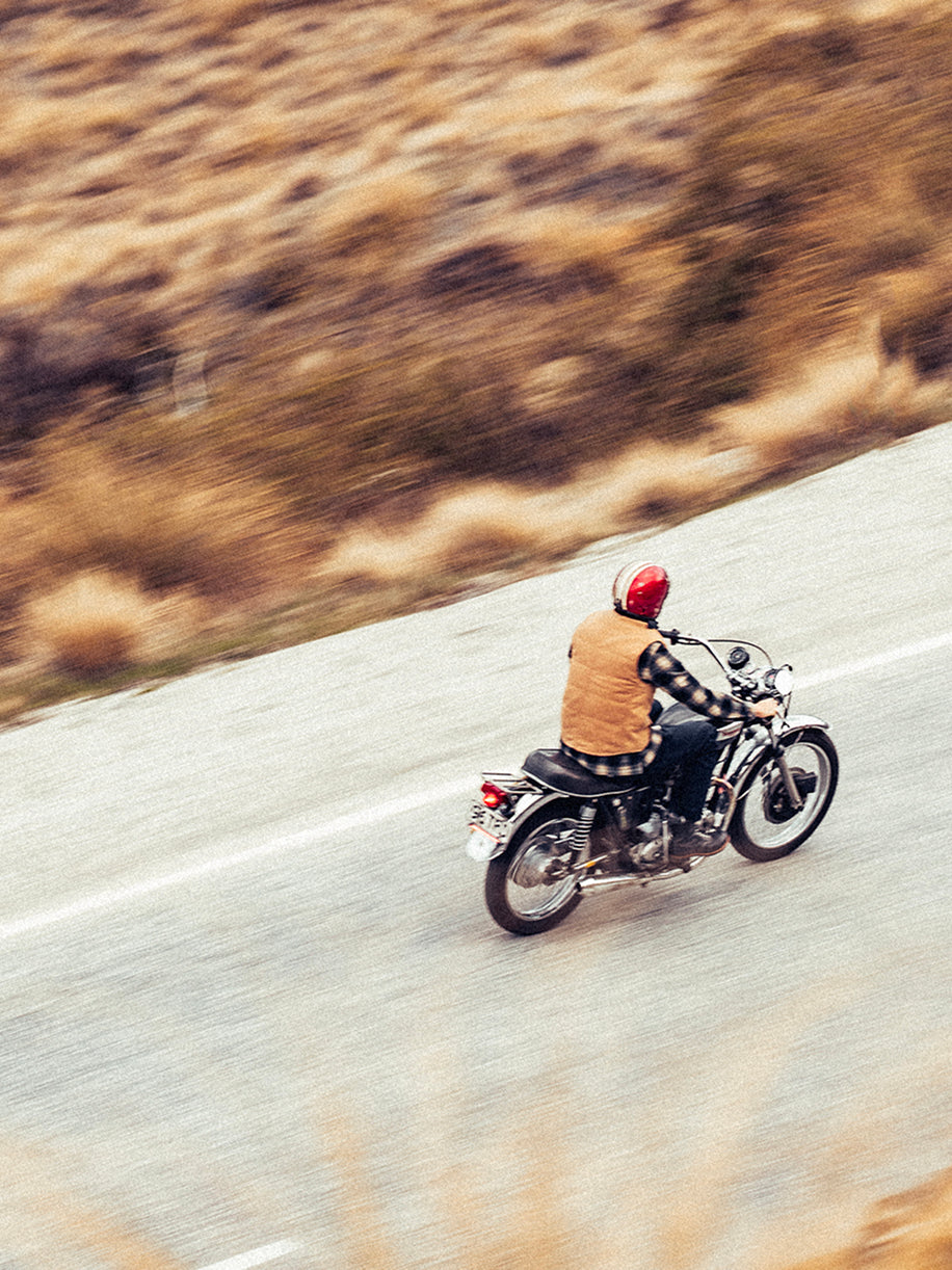 Motion-blurred action shot of motorcyclist in striped shirt and tan vest riding vintage custom motorcycle on open road with golden hillside in background