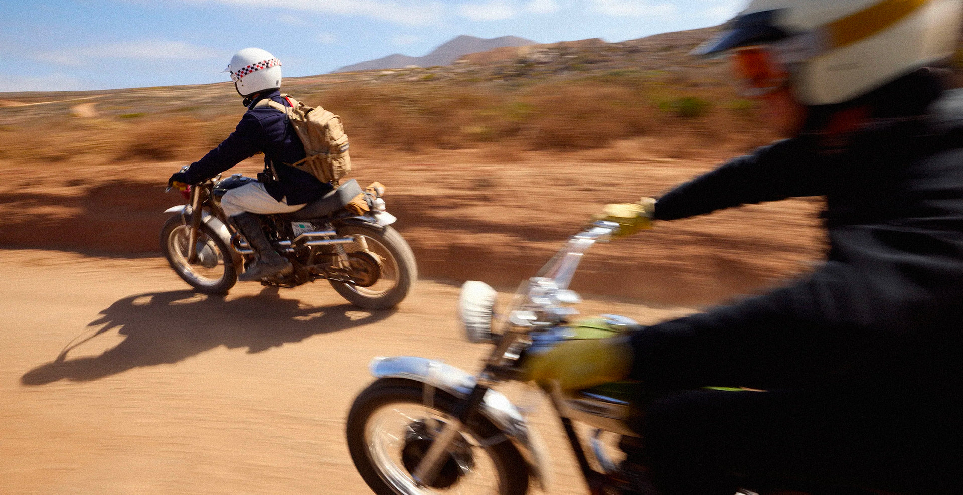 Two people riding dirt bikes on a desert track with mountains in the background.