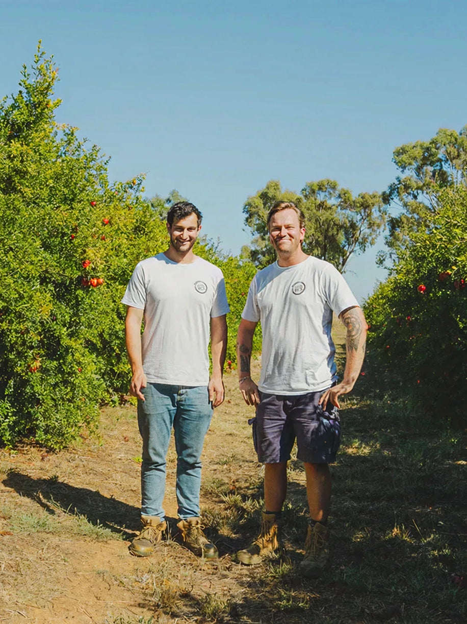 Two farmers standing in sunny orchard wearing matching gray t-shirts with circular logo, surrounded by fruit trees and green foliage