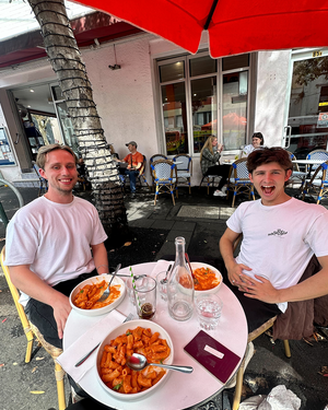 Two men dining at outdoor cafe table under red umbrella with large plates of pasta and bottle of water