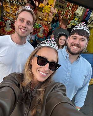 Group selfie at carnival with colorful stuffed animal prizes in background, people wearing novelty tiaras and sunglasses