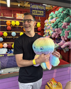 Person in black t-shirt and glasses at carnival game booth holding large rainbow gradient plush octopus toy with colorful prizes displayed behind
