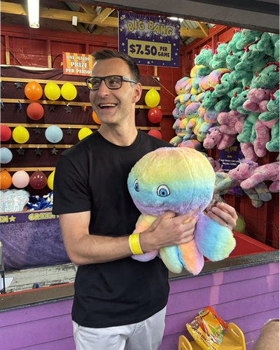Person in black t-shirt and glasses at carnival game booth holding large rainbow gradient plush octopus toy with colorful prizes displayed behind
