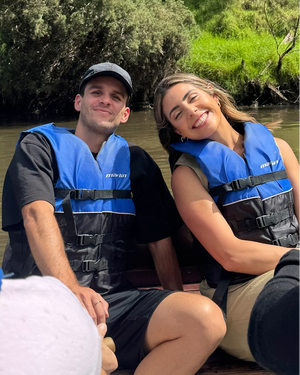 Two people in blue and black life jackets during outdoor water activity with green foliage in background