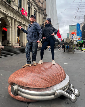 Two people posing on oversized sculpture in city plaza near historic architecture