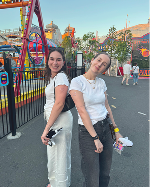 Two people posing back-to-back at amusement park at golden hour with rides visible in background