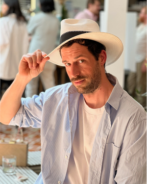 Man tipping wide-brim straw fedora hat wearing light blue striped shirt at outdoor daytime event