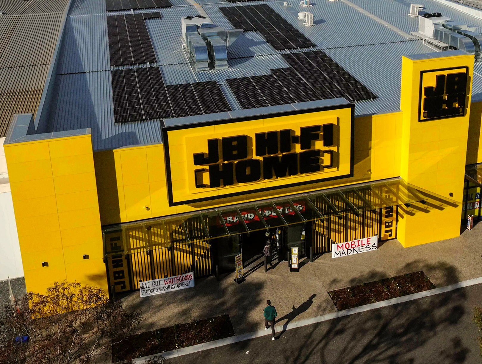 Aerial view of JB Hi-Fi retail store exterior with iconic yellow and black signage, solar panels on roof, and promotional banners at entrance
