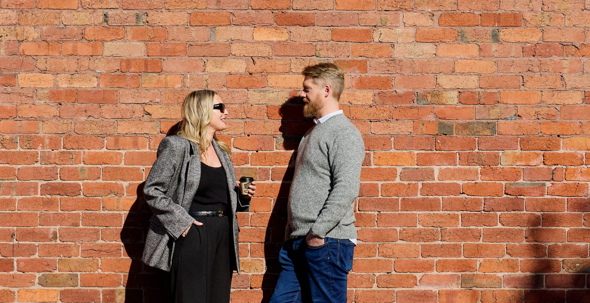 Two people standing against a brick wall, engaged in conversation.