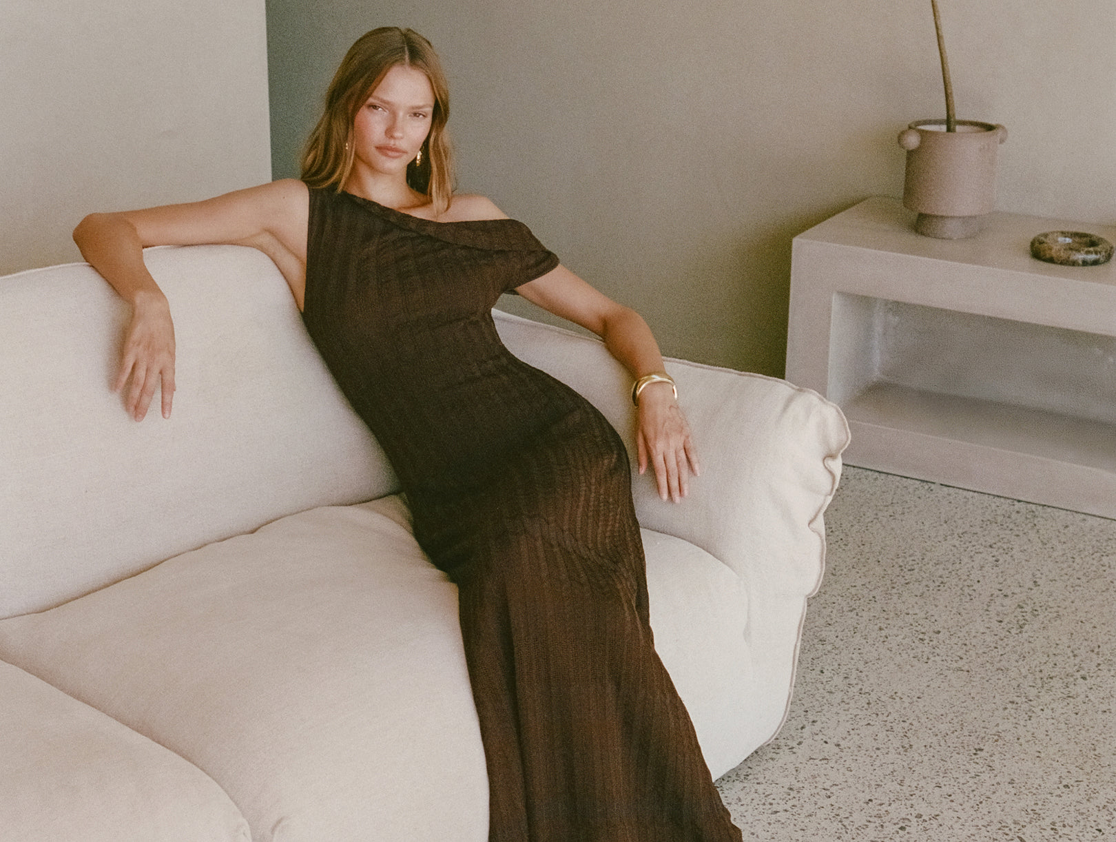 Woman in elegant olive green ribbed knit maxi dress with gold bangles reclining on white sofa in minimalist modern interior