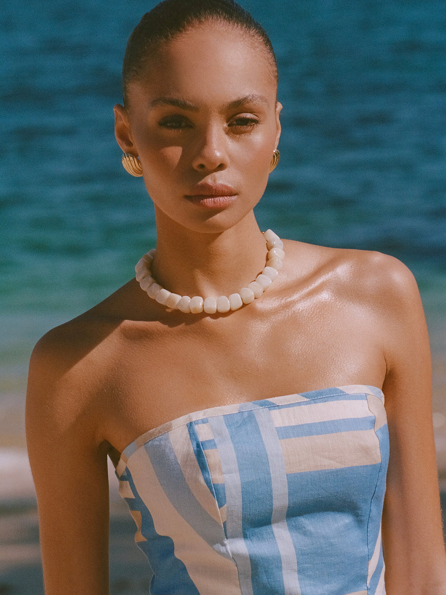 Woman with slicked back hair wearing white beaded choker necklace and blue striped bandeau top with ocean backdrop