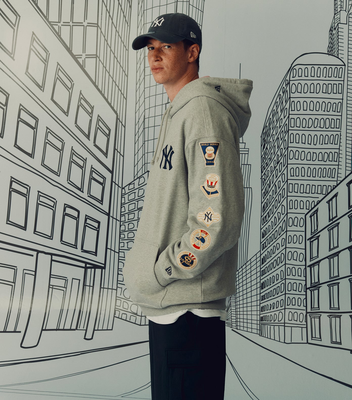 Person wearing a gray hoodie with patches in front of a cityscape illustration